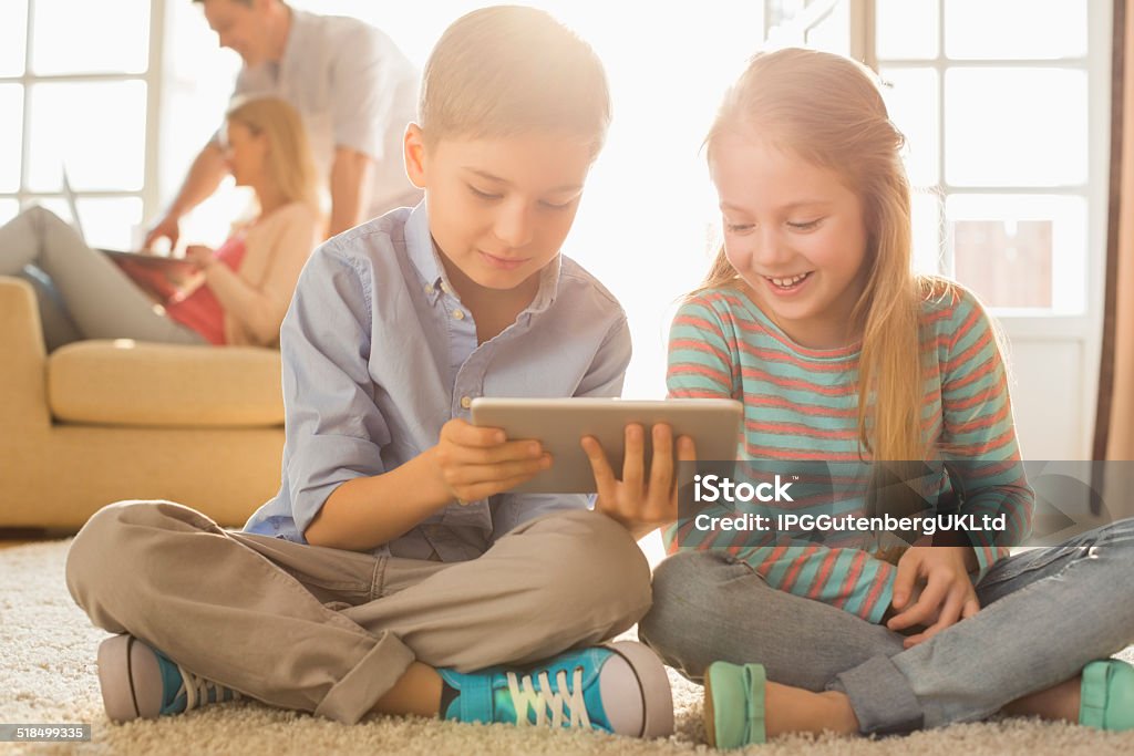 Happy siblings using digital tablet on floor Happy siblings using digital tablet on floor with parents in background 8-9 Years Stock Photo