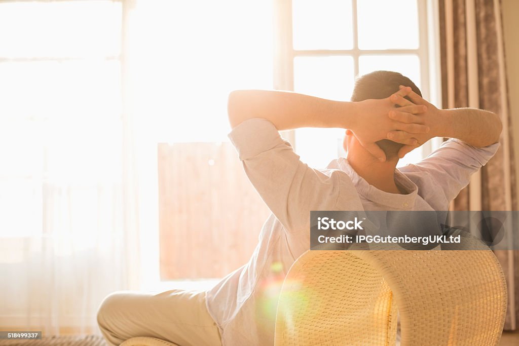 Rear view of man relaxing on chair at home Hands Behind Head Stock Photo