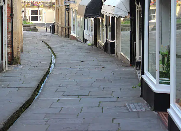 Photo showing medieval shops and houses lining the pedestrianised, flagstone shopping street of Cheap Street, located in the historic market town of Frome, Somerset, England.  This thoroughfare is particularly unusual, as it has a narrow stream / rill running through the centre of the street, fed by a nearby natural street at the top of the hill.