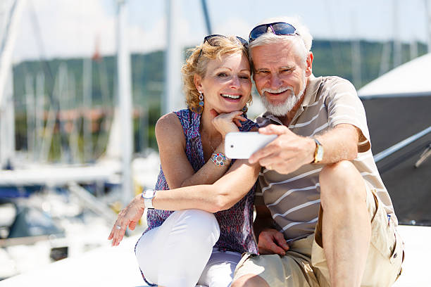 Senior couple having fun with smart phone on yacht The image shows a senior couple sailing and having fun on a yacht. They are sitting on a yacht, smiling and taking a selfie with a smartphone. In the background we can see the harbor and some boats. yacht selfie stock pictures, royalty-free photos & images