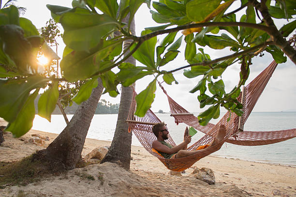 Man enjoying vacations on hammock with tablet Young man relaxing on a hammock on a tropical island using a digital tablet. hammock men lying down digital tablet stock pictures, royalty-free photos & images