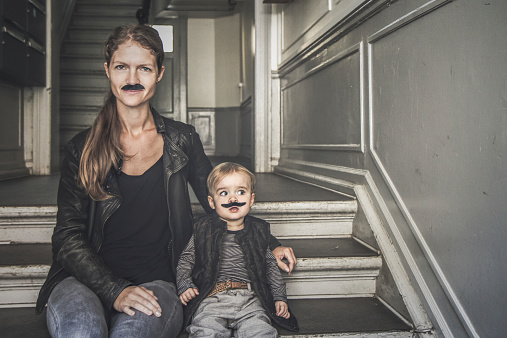 Movember: Mum and little son sitting on a staircase while looking into camera. They both wear an artificial mustache.