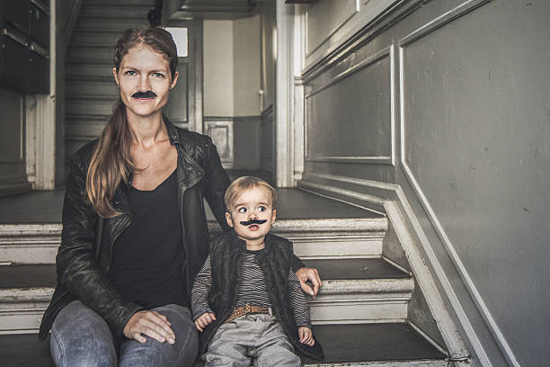 Mummys mustache Movember: Mum and little son sitting on a staircase while looking into camera. They both wear an artificial mustache. women movember mustache facial hair stock pictures, royalty-free photos & images