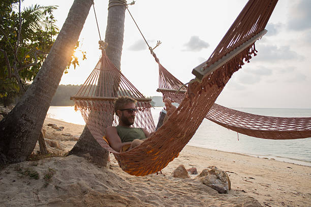 Man on hammock on tropical beach at sunset Young man relaxing on a hammock on a tropical island using a digital tablet. hammock men lying down digital tablet stock pictures, royalty-free photos & images