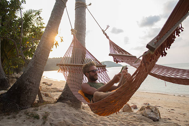 Man on tropical beach reading Young man relaxing on a hammock on a tropical island using a digital tablet. hammock men lying down digital tablet stock pictures, royalty-free photos & images