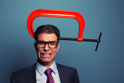 A businessman dressed in suit and wearing glasses is not responding well to business pressure. The c clamp on his head is causing him to grimace under lots of pressure. 