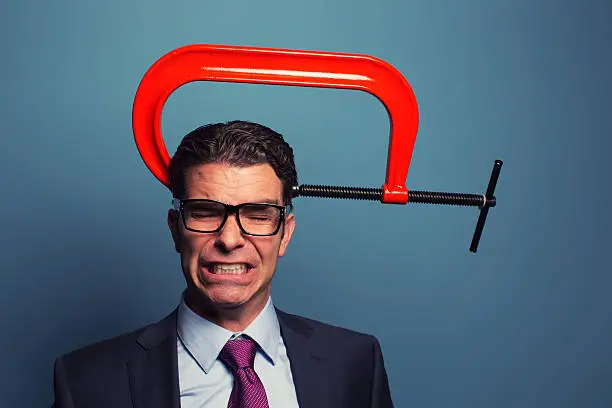 A businessman dressed in suit and wearing glasses is not responding well to business pressure. The c clamp on his head is causing him to grimace under lots of pressure. 