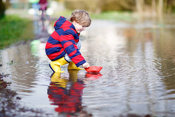 kleines kind sohn spielen mit papier-schiff in pfütze - puddle rain boot water stock-fotos und bilder