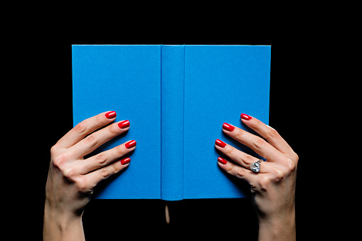 Female hands with red nail polish, reading blue hardcover book. Black background, no face