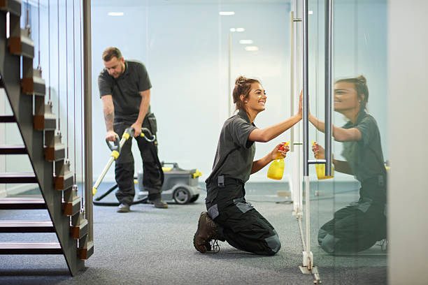 commercial cleaning contractors a female cleaning contractor is polishing the glass partition offices whilst In the background a male colleague steam cleans an office carpet in a empty office in between tenants.  .The female is smiling . cleaning equipment photos stock pictures, royalty-free photos & images