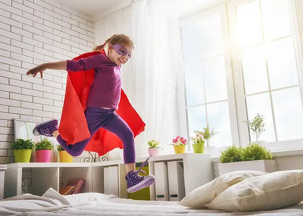 Child girl in an Superman's costume plays. The child having fun and jumping on the bed.