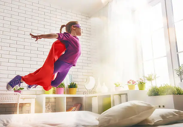 Child girl in Superhero's costume plays. The child having fun and jumping on the bed.