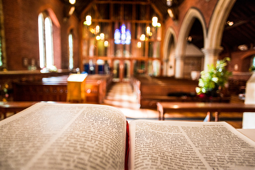 Bible open at the book of John inside an Anglican Church of England church. The focus is on the foreground of the bible, while in the background, defocused, are the pews and arches of the historic eighteenth century building. Horizontal colour image with copy space.
