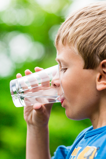 Close-up of young scandinavian child drinking fresh and pure tap water from glass with a blurred green background.