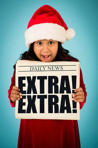 Color image of a young girl, who is wearing a Santa hat,  holding up a newspaper and making a happy/excited face.