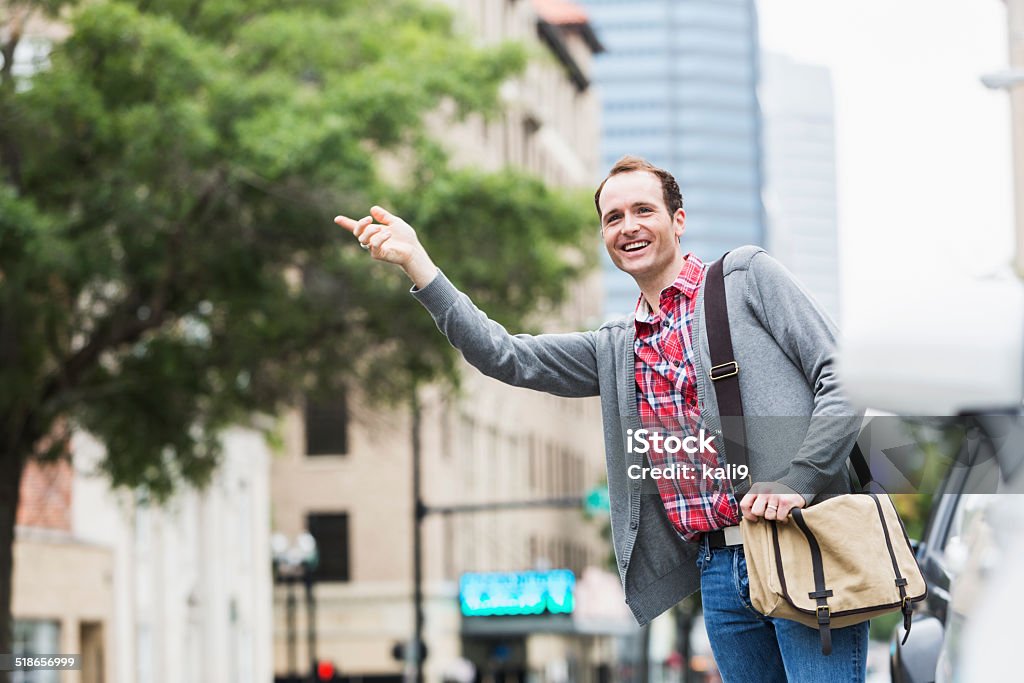 Man signaling for taxi cab A young man in the city, hailing a taxi.  He is standing next to park cars, leaning out over the street, smiling with his hand raised.  He is wearing casual clithing and jeans, carrying a shoulder bag. 20-29 Years Stock Photo