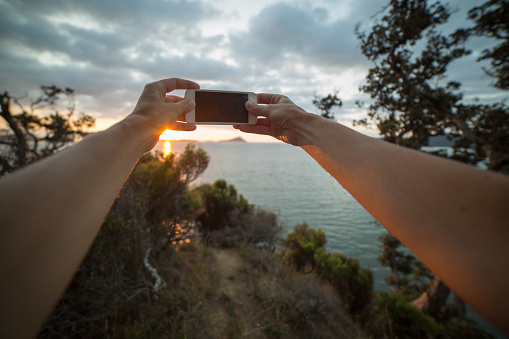 Personal perspective of a woman taking a picture of the beautiful landscape of the Hauraki Gulf at sunset using a mobile phone. Shot from Coromandel area, located in the Bay of Plenty on the North Island of New Zealand.