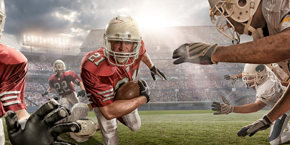 Close up, mid action image of American football player holding ball and running through rival players with teammates. The action is set on an American football field in a generic floodlit outdoor stadium under a bright and cloudy evening sky with dramatic sunlight. All players are wearing generic unbranded kit. 