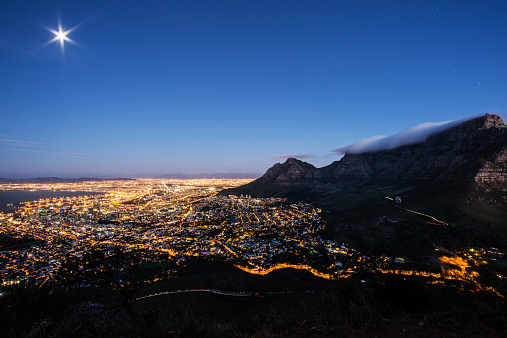 Panoramic Aerial View over illuminated Cape Town Cityscape at the foot of famous Table Mountain under clear and starry sky.