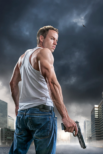 Low angle portrait image of an intense man dressed in jeans and a white vest viewed from behind turning to look towards camera, holding a generic gun.  The man in standing outdoors in a generic city with modern high rise buildings near a river under stormy evening sky at sunset.