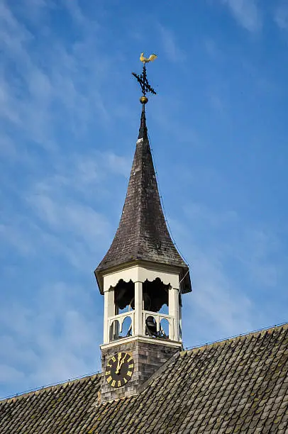 Closeup of a pointed church tower with clock, belfry, a cross and a golden weathervane dating from 1725 against the blue sky.