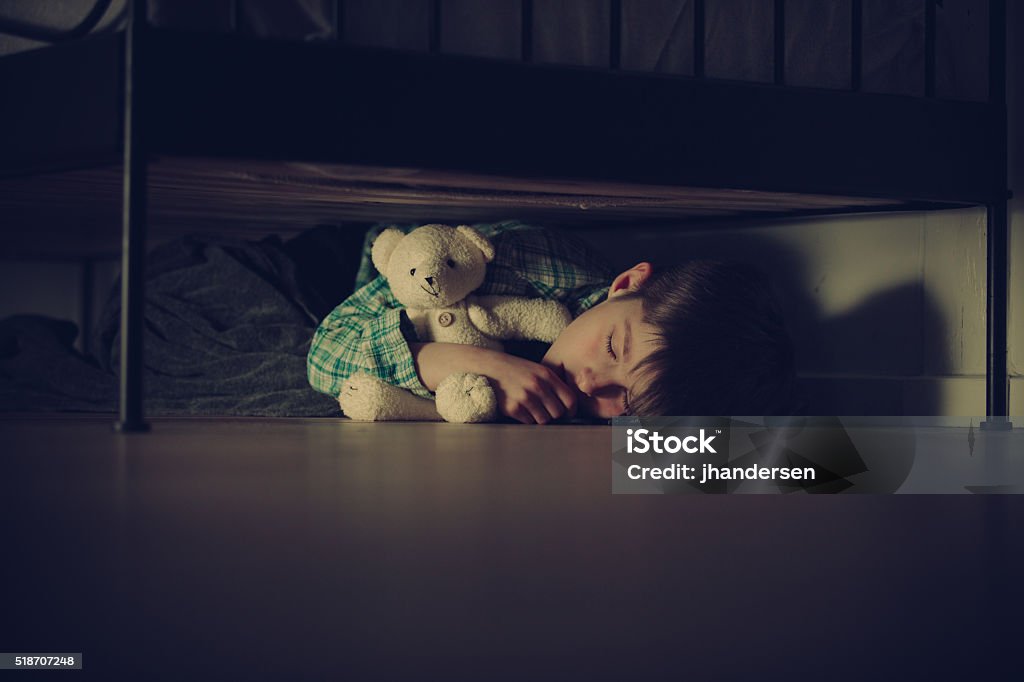 Scared Boy Sleeping Under his Bed with Teddy Bear Scared Young Boy Sleeping Under his Bed Inside his Room While Hugging his Teddy Bear Stuffed Toy at Night. Child Stock Photo
