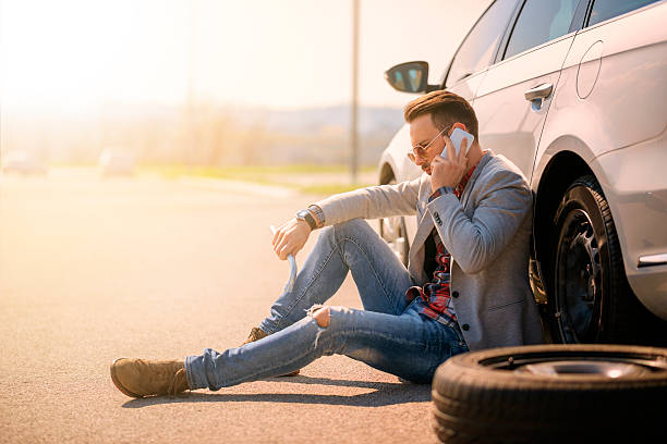 Car breakdown A young man with a silver car that broke down on the road.He is waiting for the technician to arrive. flat tire stock pictures, royalty-free photos & images