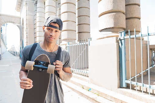 This is a horizontal, color, royalty free stock photograph of an athletic, confident, young Black man in his 20s from Brooklyn. He takes his skateboard across the Manhattan Bridge in New York City on a hot summer day. He holds his skate deck in hands and looks at the camera while listening to music with headphones. Photographed with a Nikon D800 DSLR camera.