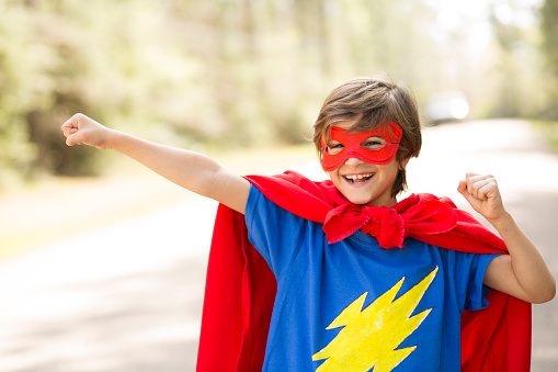 Latin descent, cute little boy plays superhero outdoors on his neighborhood street in spring or summer season.  He has made a costume for himself with red mask and 