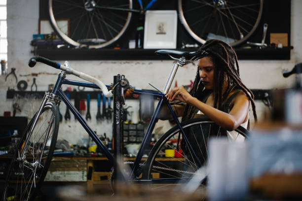 Afro woman bicycle mechanic recpairing a bike in her workshop Afro-american craftswoman repairing a bicycle in her bike repair workshop with tools and spares around her bike workshop stock pictures, royalty-free photos & images