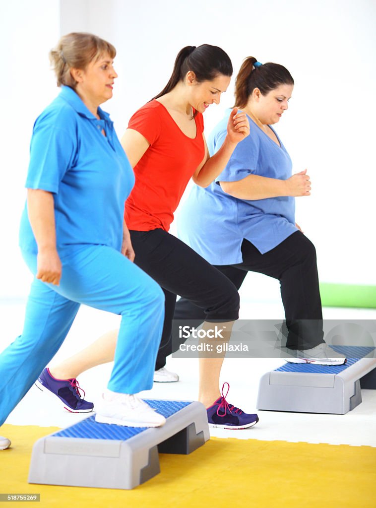 Weight loss exercise. Side view of couple of overweight  senior women doing stepper exercises with female instructor. They are standing side by side with the instructor in the middle and putting each leg on the stepper, one by one. Wearing blue sport clothes and white shoes. 40-49 Years Stock Photo