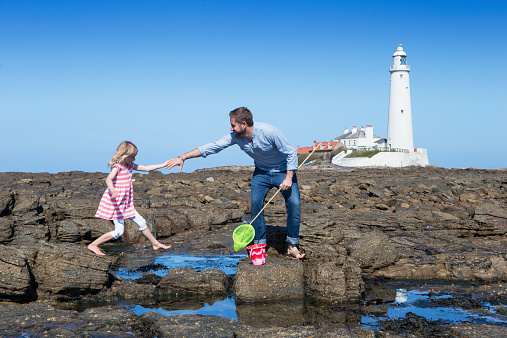 A young father helps his daughter across the rocks along the coast as they play by the rock pools. St Mary's lighthouse is in the background.