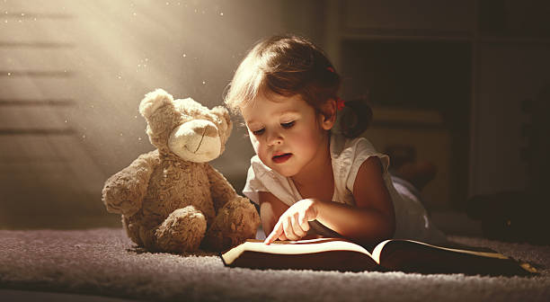 niño niña pequeña leyendo un libro de magia en la oscuridad hogar - muñeco de peluche fotografías e imágenes de stock