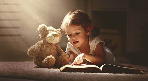 child little girl reading a magic book in dark home - speelgoedbeest stockfoto's en -beelden