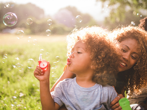 Soft glowing sunny moment between a young african-american mother and her cute curly haired baby boy blowing colourful bubbles on the meadow.