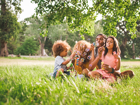 Cute african-american family sitting in a gorgeous picturesque park while playing with their kids and blowing bubbles on a beautifull sunny afternoon