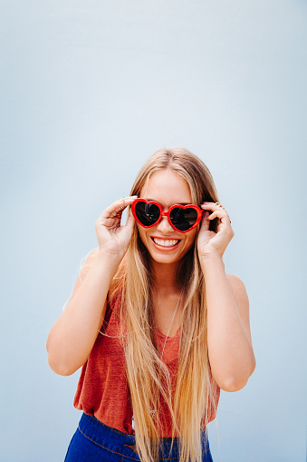 Beautiful young blond woman with sunglasses with a hearth shape, isolated on blue wall background