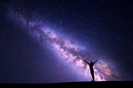 Landscape with colorful purple Milky Way. Night sky with stars and silhouette of a standing sporty girl with raised-up arms on the hill. Beautiful Universe. Space background