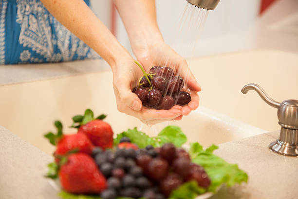 Woman rinses red grape fruits in home kitchen sink. Unrecognizable woman washes, rinses fresh red grapes in home kitchen sink. Water sprays from faucet onto the juicy fruit. A plate of organic fruit in foreground includes: strawberries, blueberries, and grapes. She is preparing a healthy snack or meal. Handful of Grapes stock pictures, royalty-free photos & images