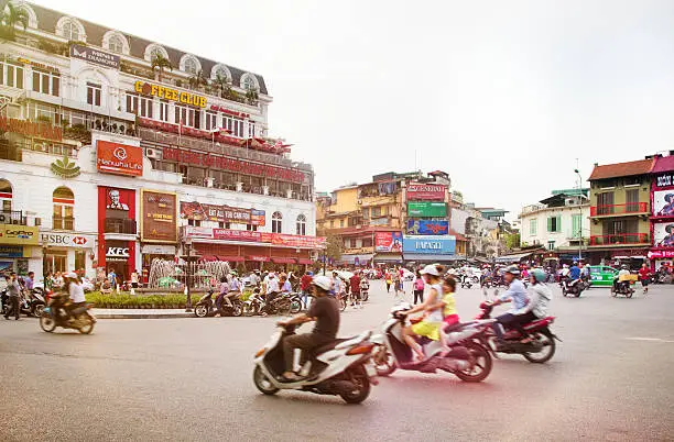 Busy Hanoi Fountain roundabout with traffic and multiple businesses aimed at tourists. Sveral people are using the roundabout on their scooters, while others are walking or stopped, talking to each other.