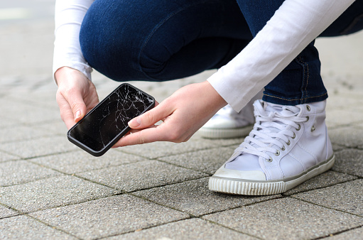 Close up view on kneeling person in jeans and shoes picking up broken phone on stone paved sidewalk outdoors