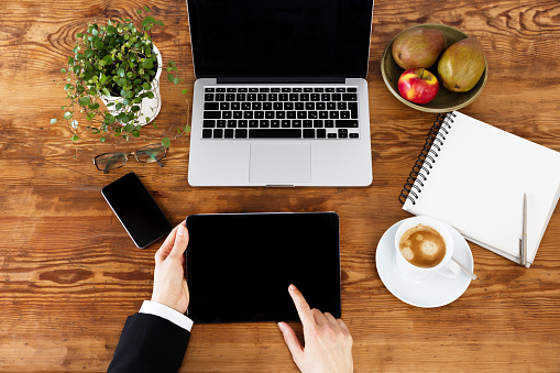 Business Desk – Top View Of Woman Hands Using Tablet, Laptop And Smartphone Sitting By The Wooden Table Surrounded With Coffee, Plant And Watermelon