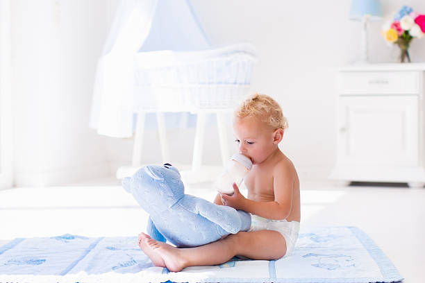 Cute blond curly baby boy wearing a diaper drinking milk on a sunny morning playing with his toy teddy bear in sunny white bedroom. Kids drink formula. Toddler with plastic bottle. Child nutrition.