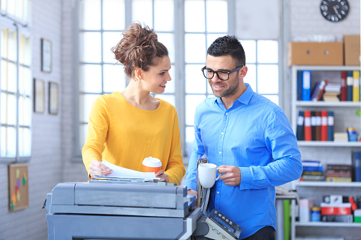 Coworkers working and drinking coffee together at the office. Woman wears yellow blouse and holding plastic cup and printed documents. Man holding cup, wears blue shirt and eyeglasses. Wall with tall windows and shelves with boxes and folders as background. A copy machine in foreground.