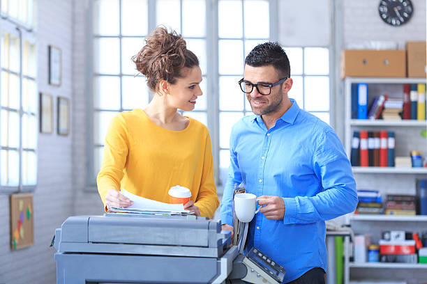 Coworkers working and drinking coffee together at the office Coworkers working and drinking coffee together at the office. Woman wears yellow blouse and holding plastic cup and printed documents. Man holding cup, wears blue shirt and eyeglasses. Wall with tall windows and shelves with boxes and folders as background. A copy machine in foreground. computer printer stock pictures, royalty-free photos & images