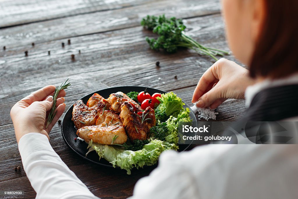 Food decoration. Chief adds spices and herbs Decoration of chicken wings on wooden table. Chief prepares grilled spicy chicken with fresh vegetables. Over the shoulder view on food decoration.  Animal Body Part Stock Photo