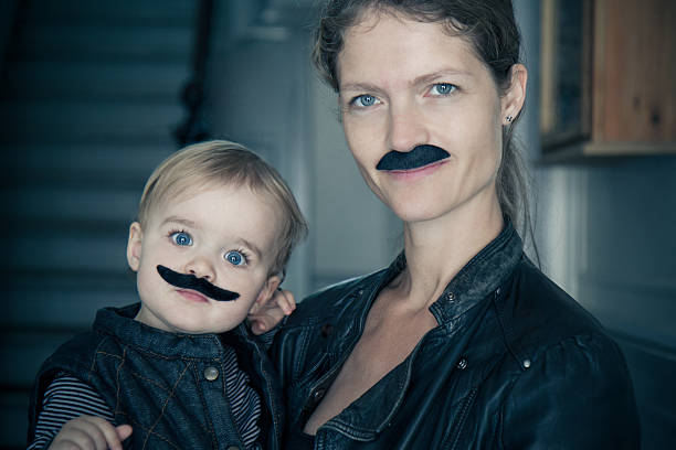 Hairy Family Movember: Mum holding her baby boy while looking into camera. They both wear an artificial mustache. women movember mustache facial hair stock pictures, royalty-free photos & images