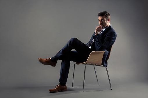 Studio portrait of an attractive adult man in a suit sitting on a chair, isolated on grey background.
