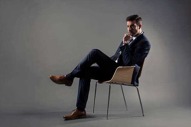 Timeless Sophistication Studio portrait of an attractive adult man in a suit sitting on a chair, isolated on grey background. legs crossed at knee stock pictures, royalty-free photos & images