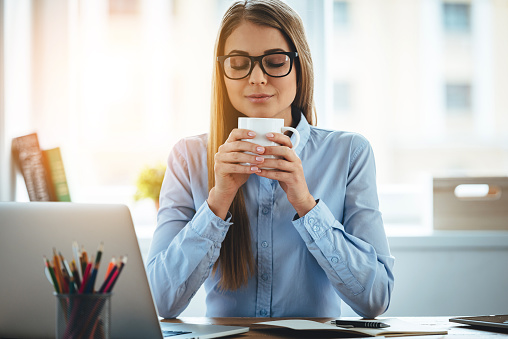 Young beautiful woman holding coffee cup and keeping eyes closed while sitting at her working place